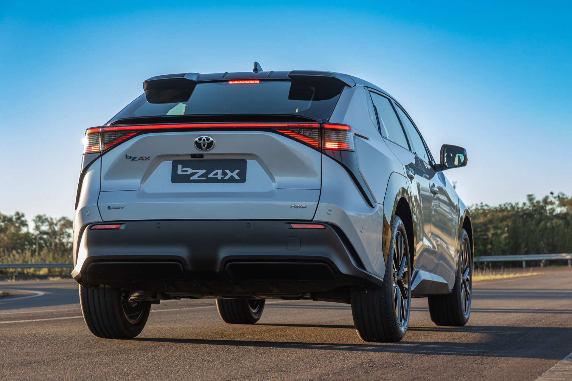 Rear view of a white Toyota bZ4X electric SUV parked on a road, showcasing its modern design and rear lights against a blue sky.