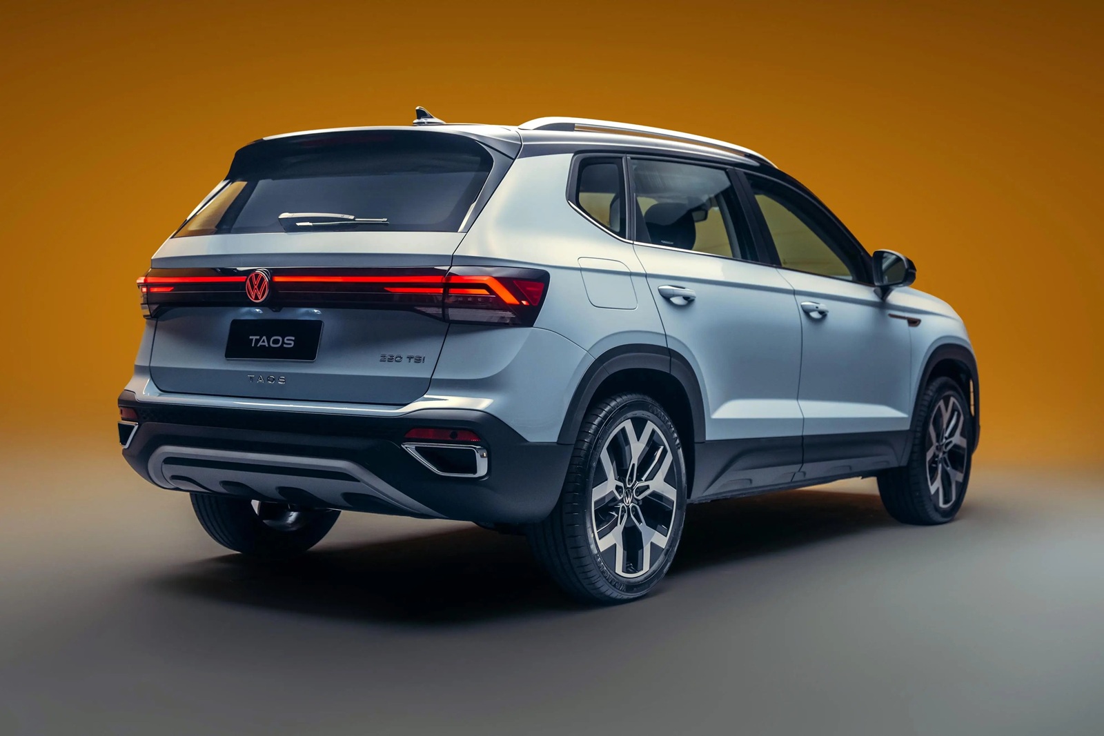 A rear view of a Volkswagen Taos SUV with a silver exterior, showcasing its sleek design and modern taillights against a gradient orange background.
