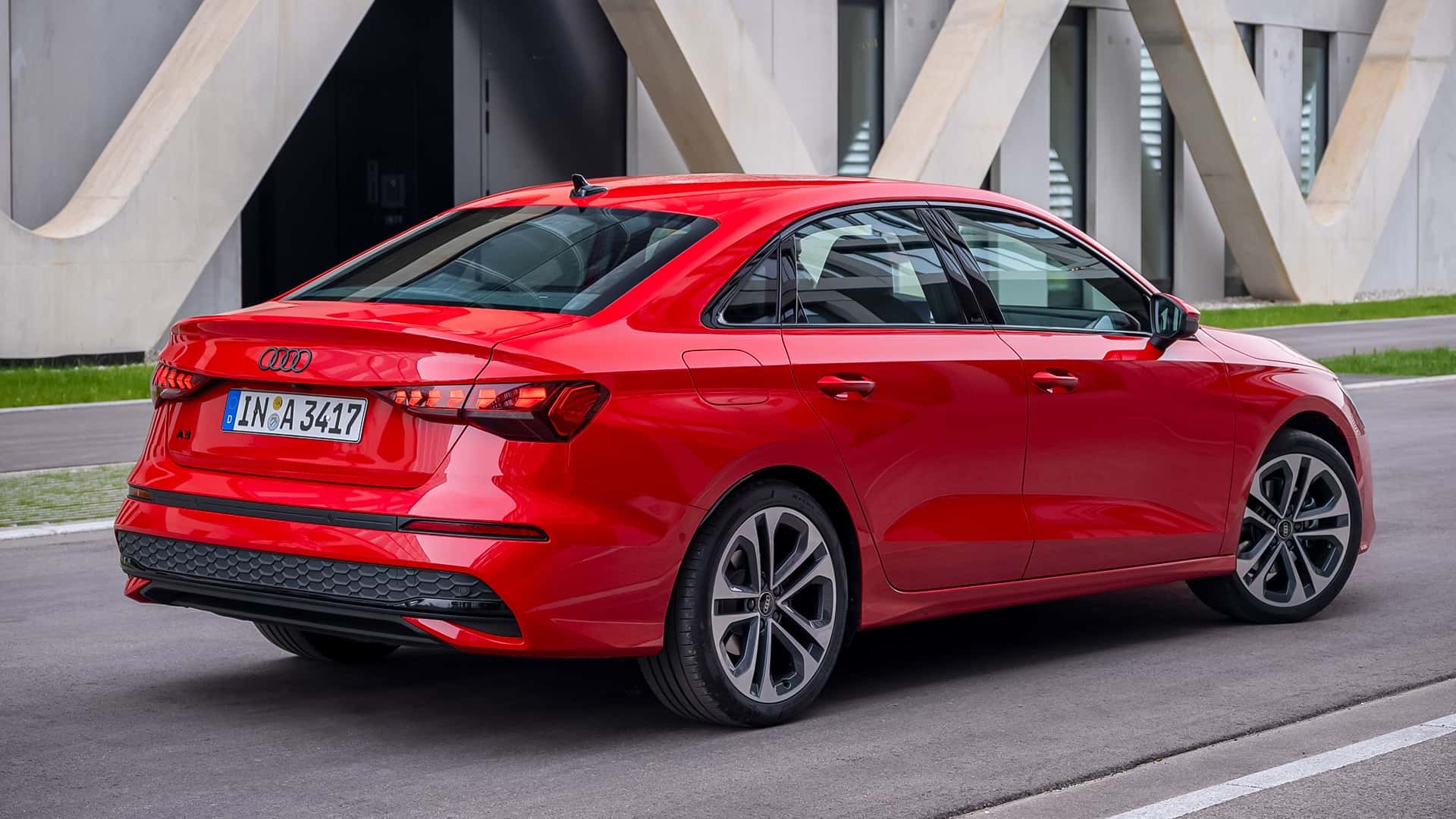A red Audi A3 sedan parked on a modern road with a contemporary building in the background.