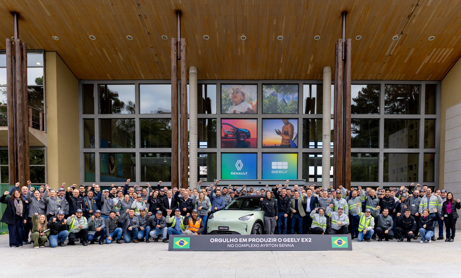 Grupo de funcionários posando em frente ao Complexo Ayrton Senna, celebrando a produção do carro Geely EX2, com o painel da empresa ao fundo.