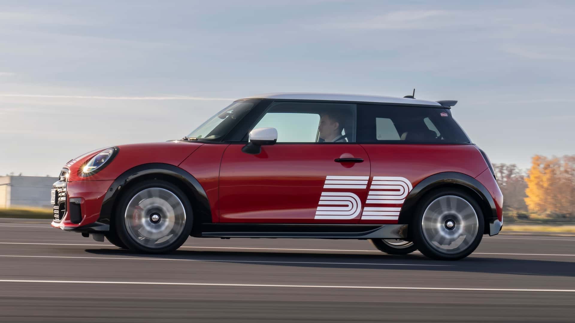 A red Mini Cooper car with white racing stripes driving on a paved road, showcasing its side profile against a clear sky.