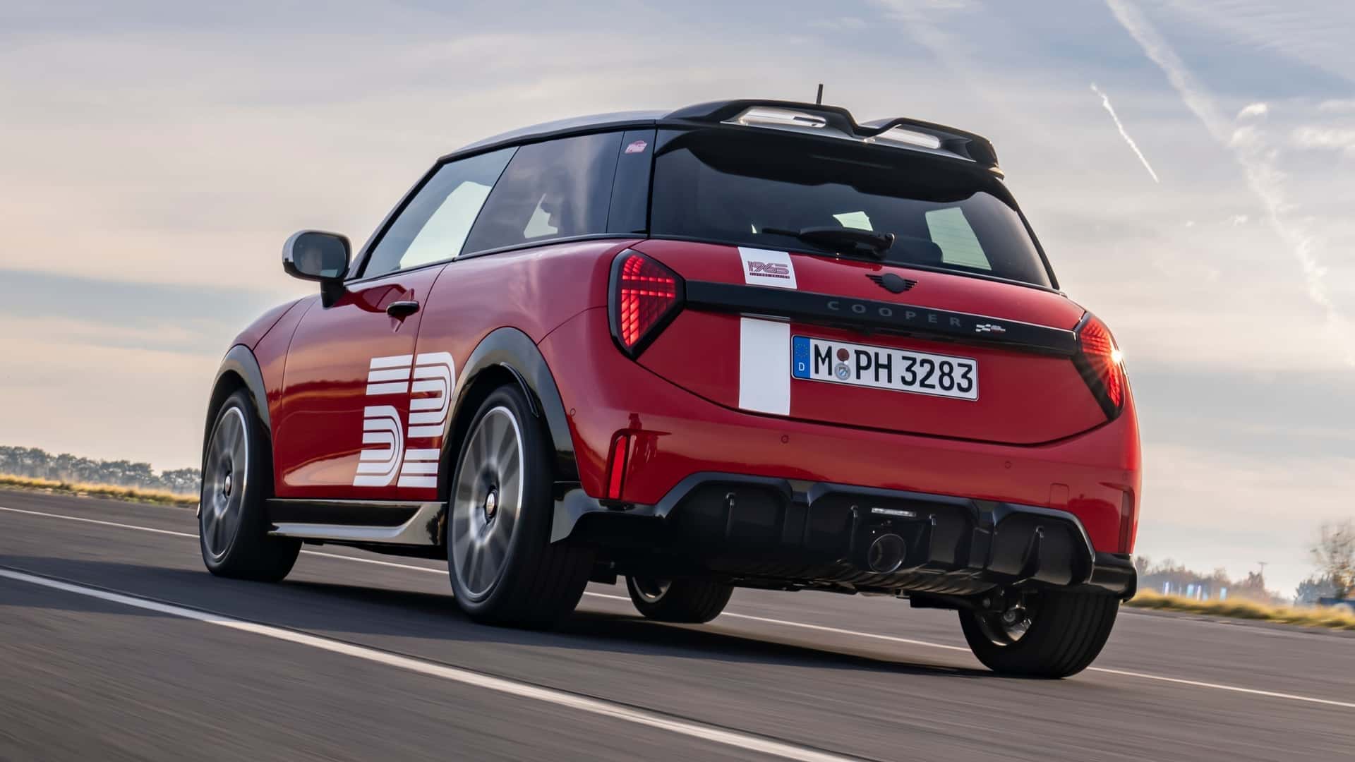 A red Mini Cooper car with a sporty design, featuring a large '52' graphic on the side, driving on an open road with a clear sky in the background.