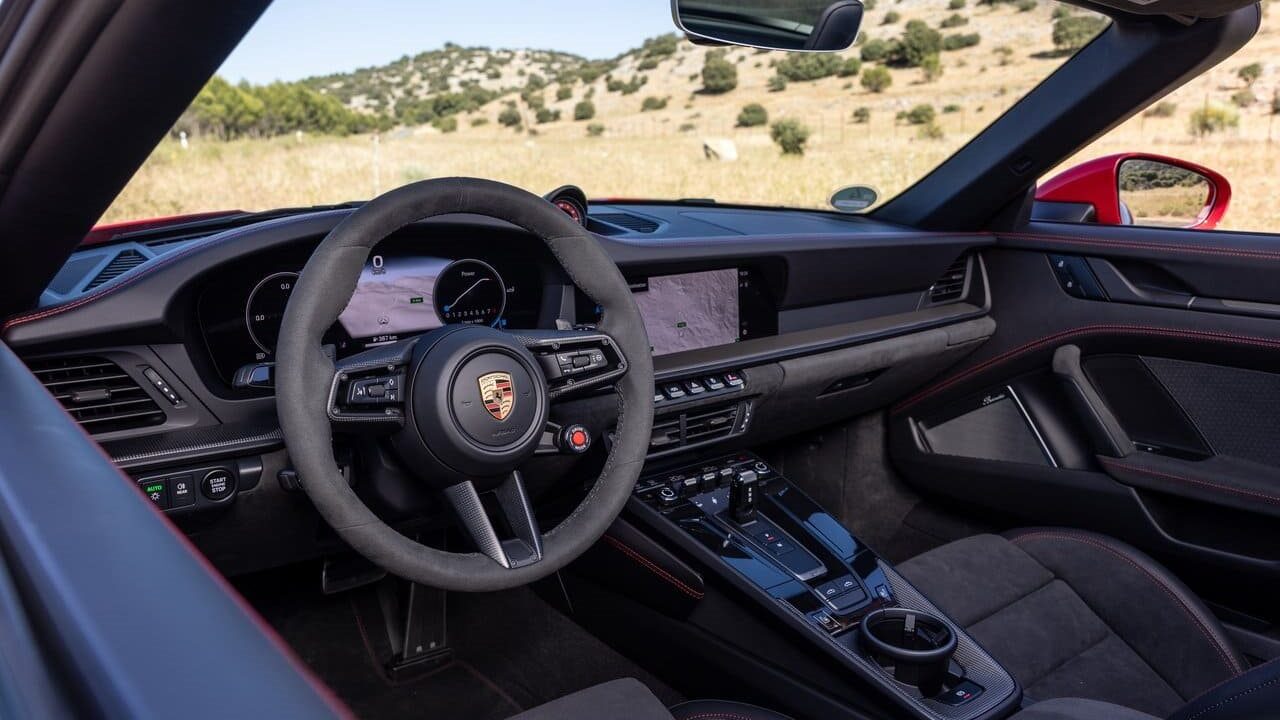 Interior view of a Porsche sports car, showcasing the steering wheel, digital dashboard display, and sleek controls set against a natural backdrop.