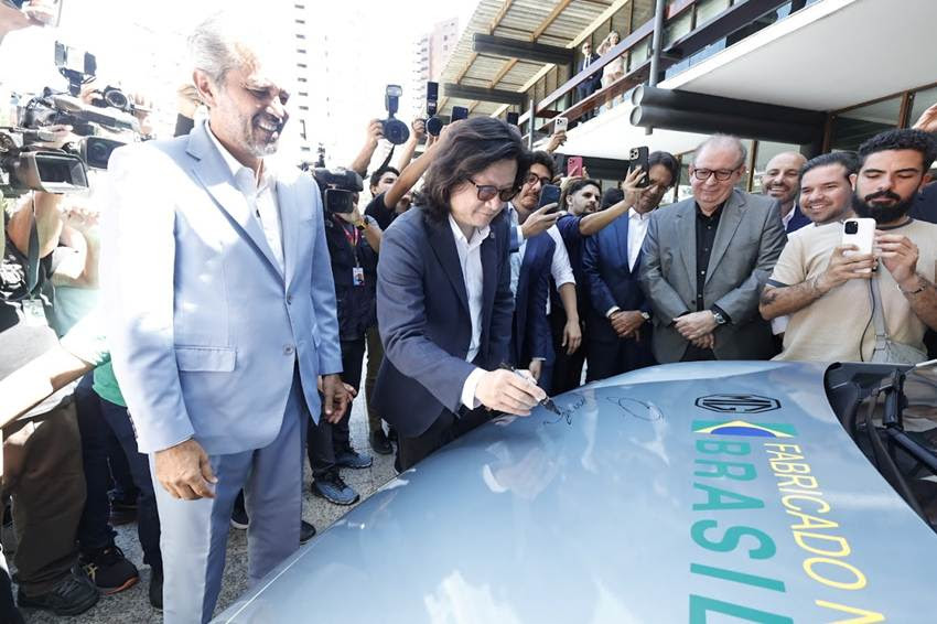 Evento de lançamento de um veículo, com pessoas observando enquanto uma figura pública assina o capô do carro.