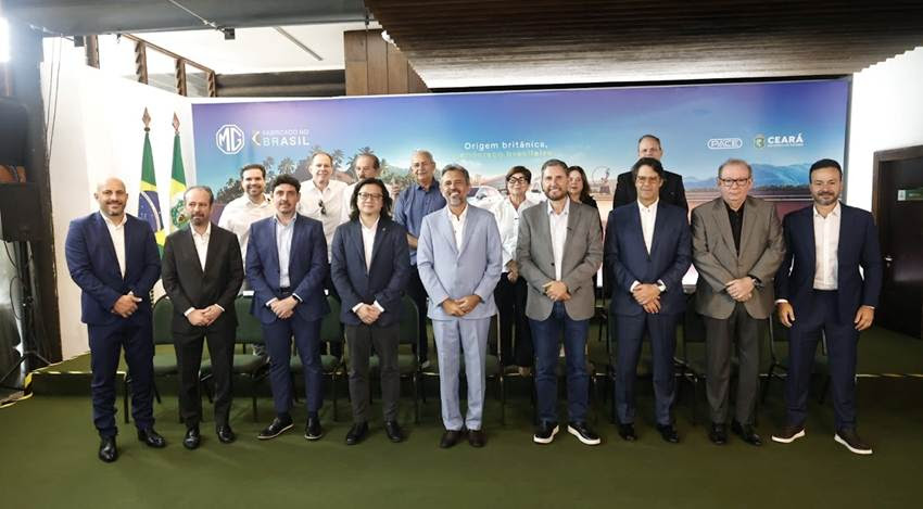 Grupo de pessoas posando para foto em evento, com bandeiras do Brasil e do Ceará ao fundo, e um banner com o texto 'Origem britânica'.