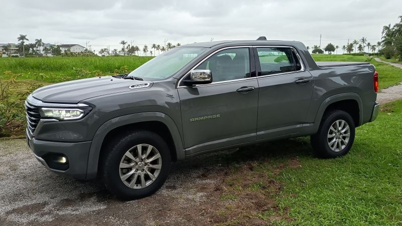 Uma caminhonete cinza estacionada em um caminho de terra, rodeada por vegetação e com um céu nublado ao fundo.
