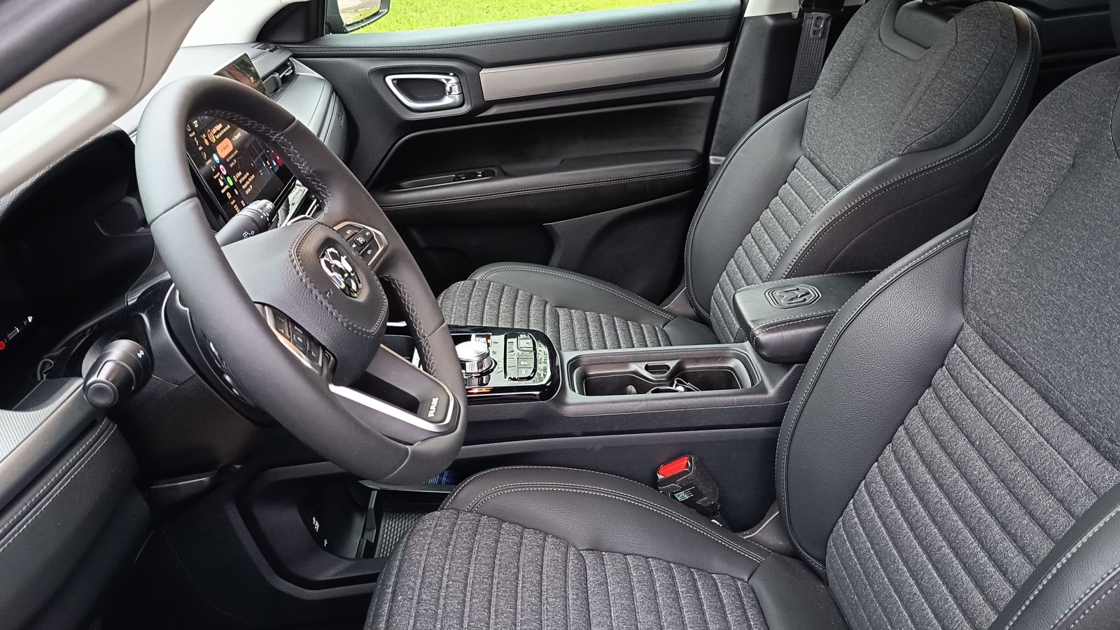 Interior de um carro, mostrando o volante e assentos em couro com detalhes em tecido, além do painel de controle com tela digital.