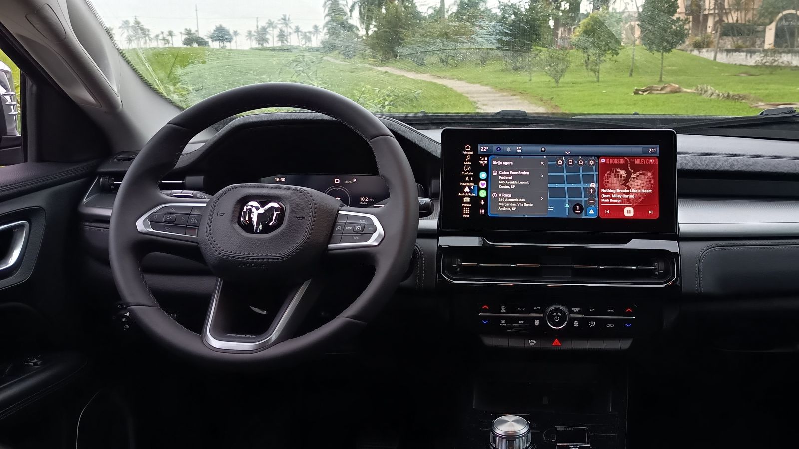 Interior de um carro com volante e painel central, apresentando uma tela multimídia e informações do painel.