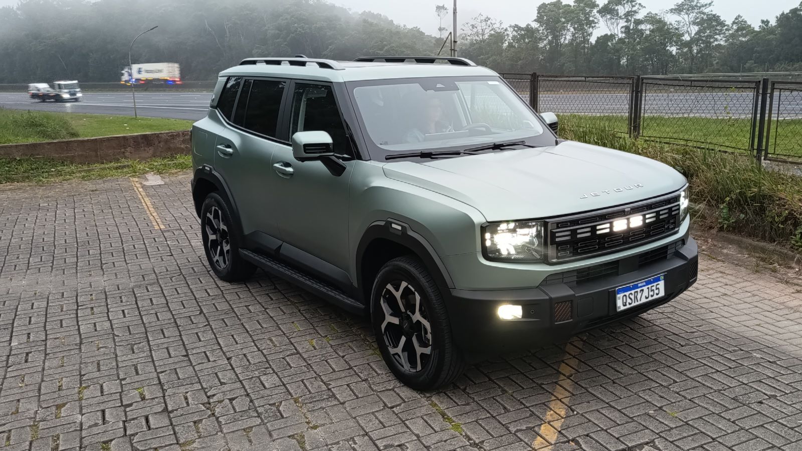 SUV moderno estacionado em um local com vegetação ao fundo e uma estrada à esquerda, com neblina no ar.