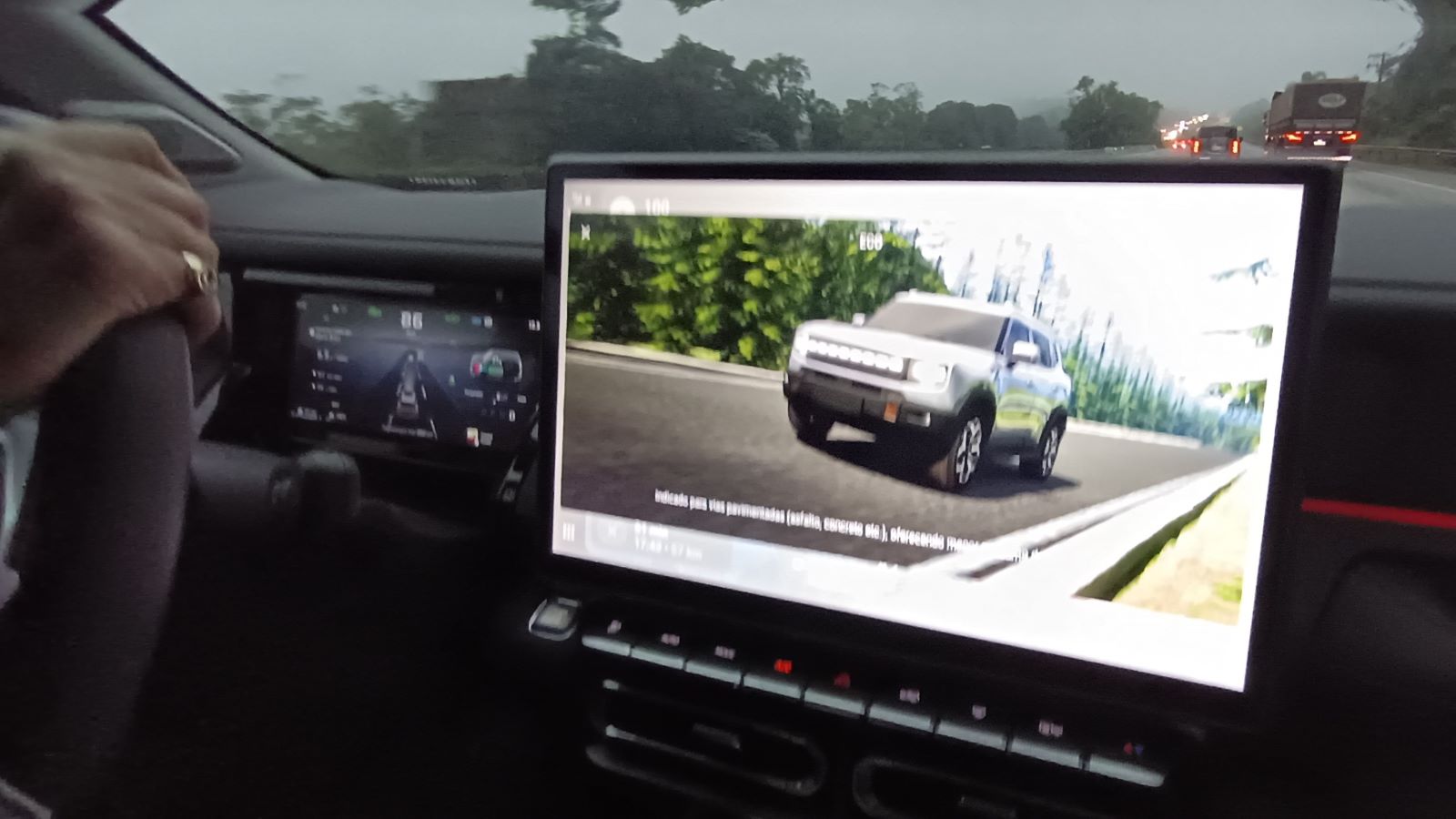 Visão do interior de um carro com uma mão no volante, mostrando uma tela digital com uma animação de um SUV em uma estrada.