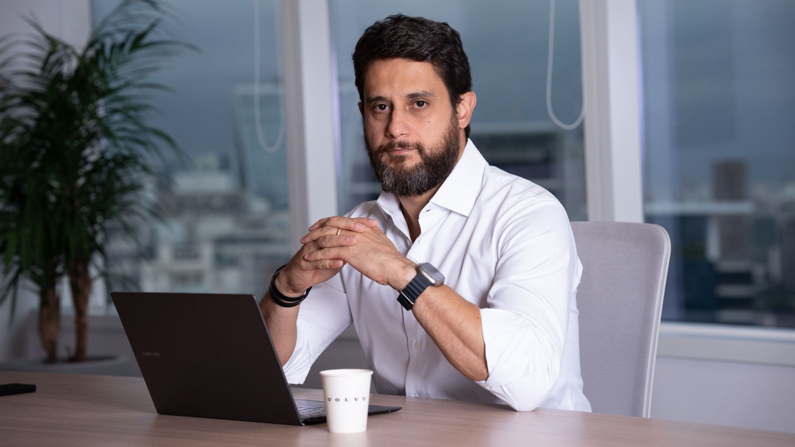 Homem com barba sentado em uma mesa de escritório, usando camisa branca, com laptop e copo de café à sua frente.