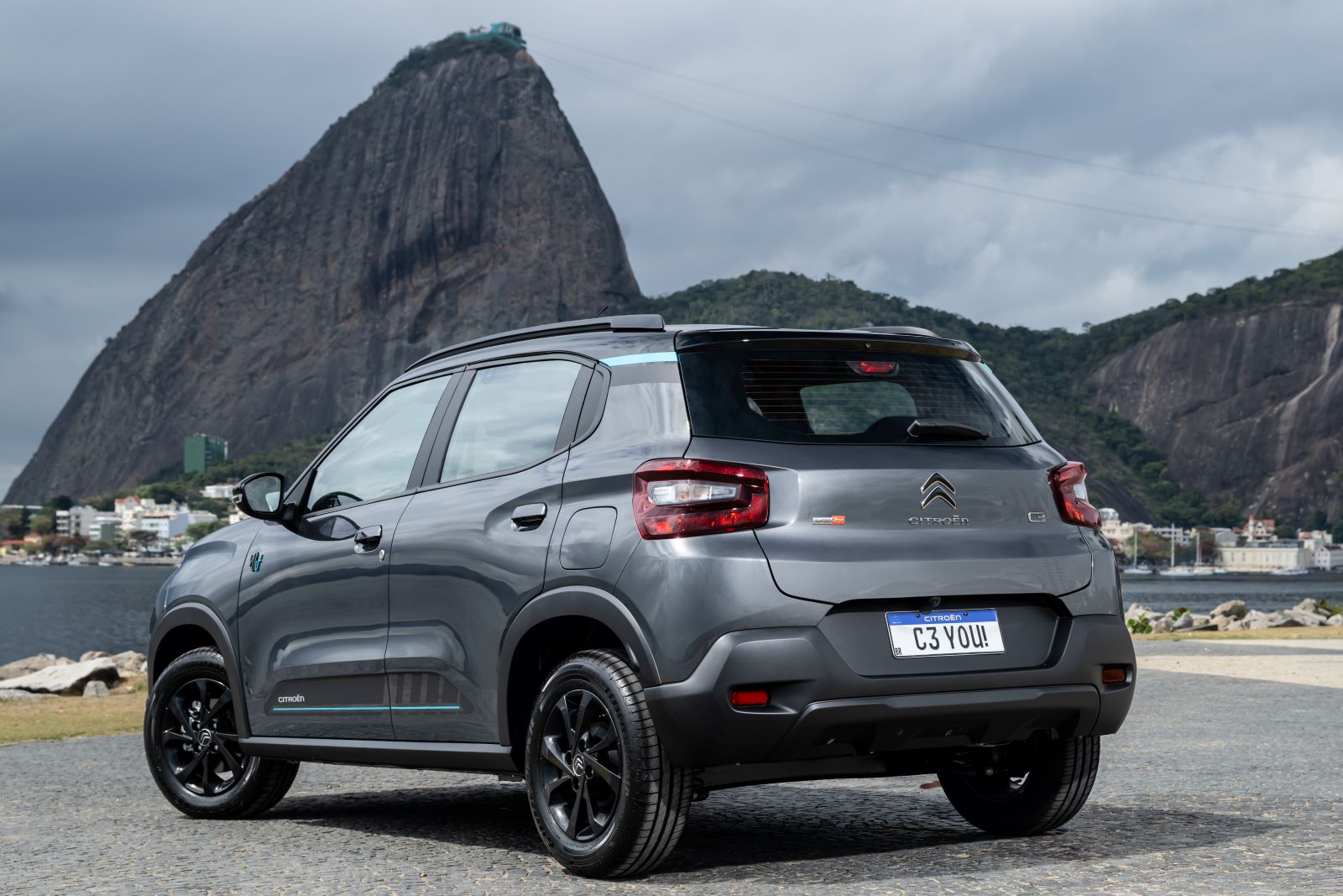 Um carro cinza da marca Citroën, modelo C3, estacionado à beira da água, com o Pão de Açúcar ao fundo em um dia nublado.